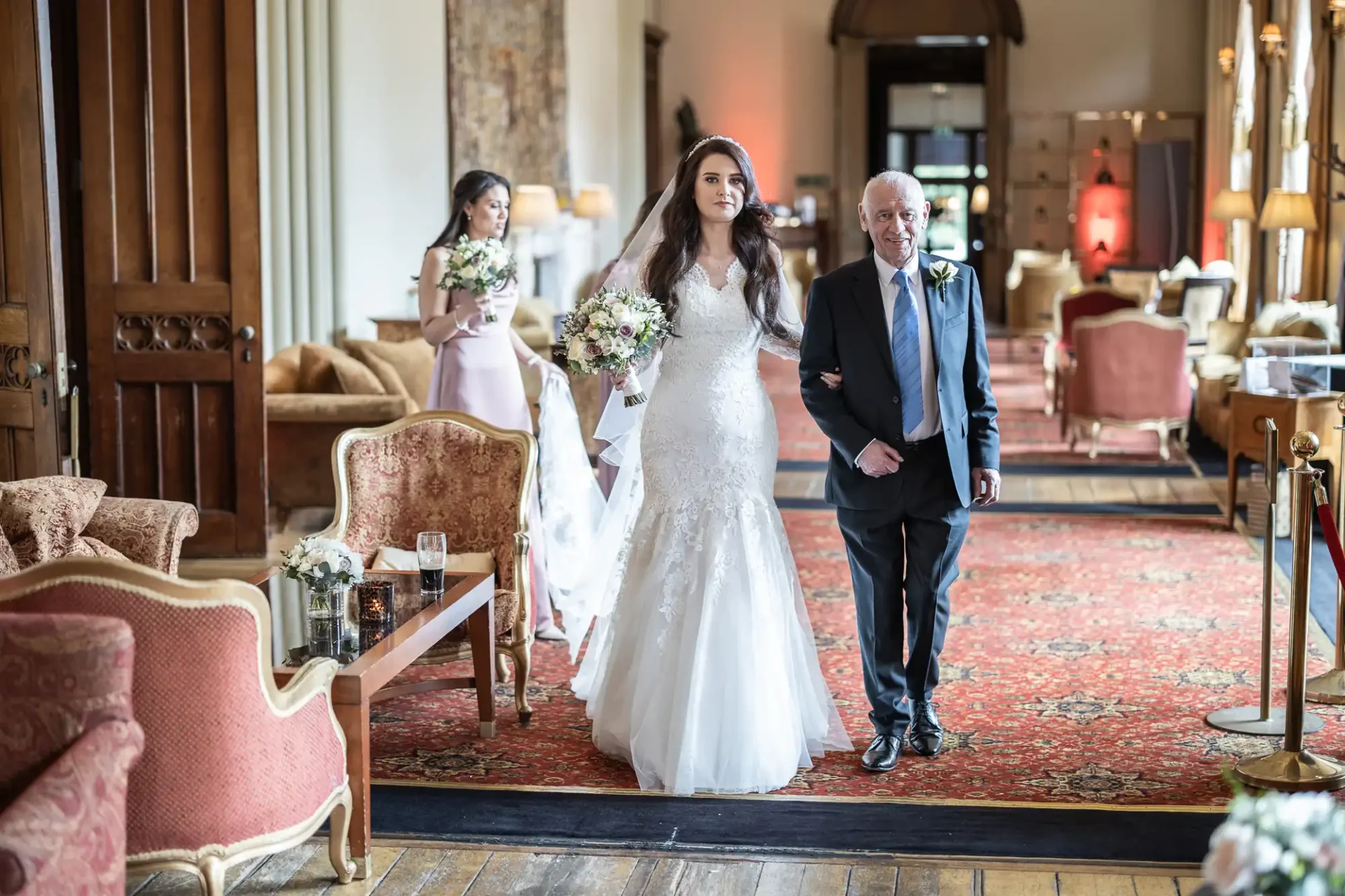 A bride in a white gown walks down the aisle with an older man in a suit, followed by a bridesmaid holding flowers, in an elegantly decorated room.