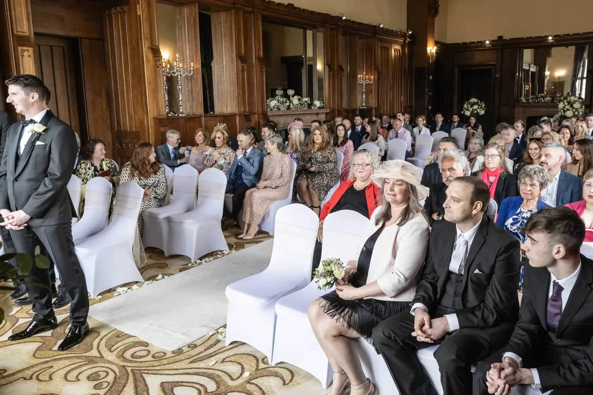 A groom stands at the front of a wedding venue facing seated guests in a wood-paneled room with white chair covers.