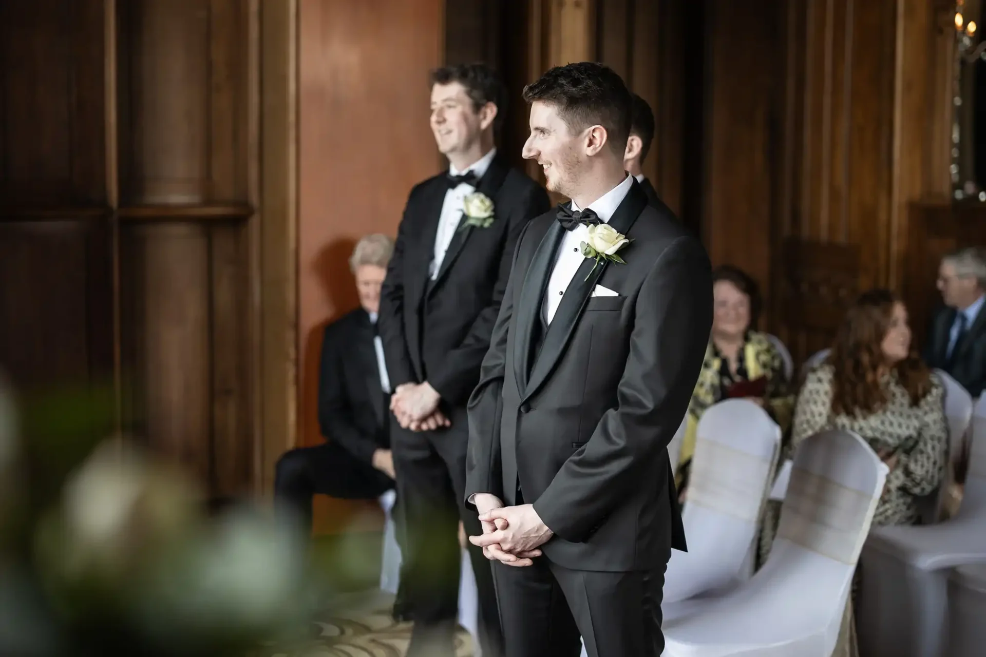 Two men in suits stand in the aisle, smiling, during a ceremony. Seated guests are in the background, and the setting appears to be a formal venue with wood paneling.