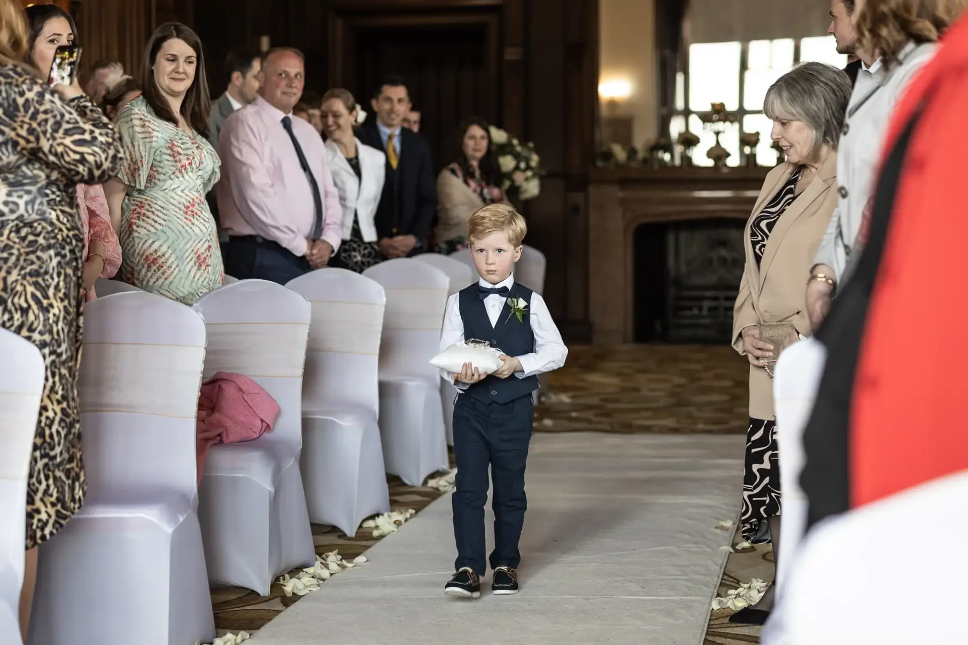 A young boy in a suit walks down an aisle with a ring pillow, surrounded by seated guests on both sides.