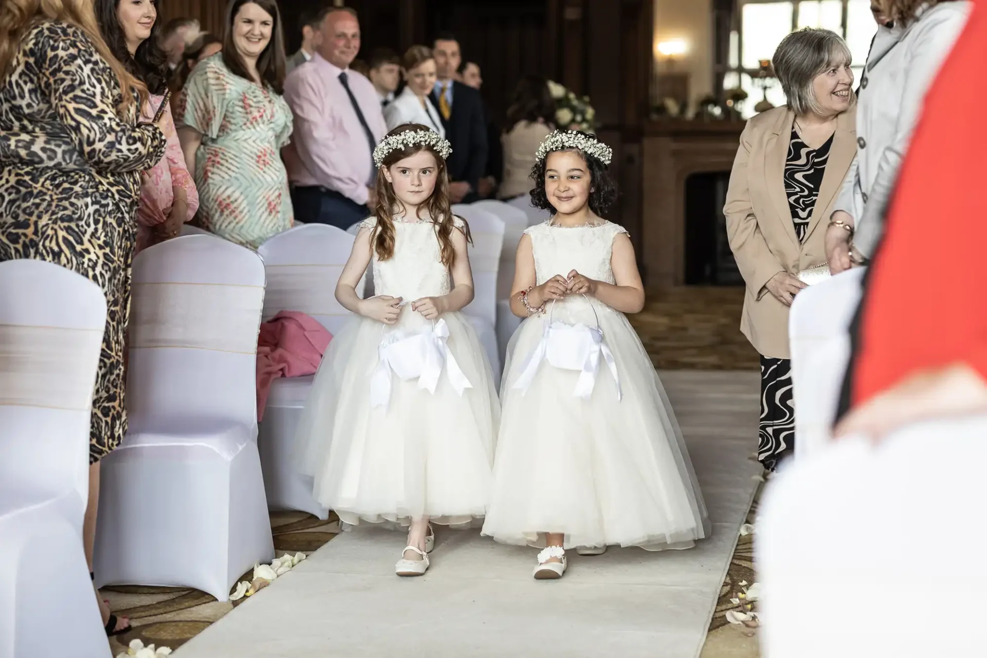 Two young flower girls in white dresses and floral headbands walk down the aisle, surrounded by seated guests.