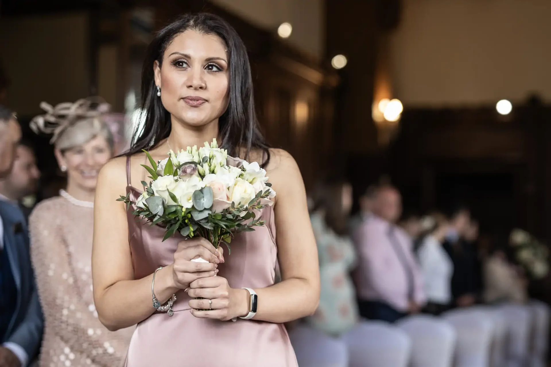 Woman holding a bouquet standing in a room with seated people in the background.