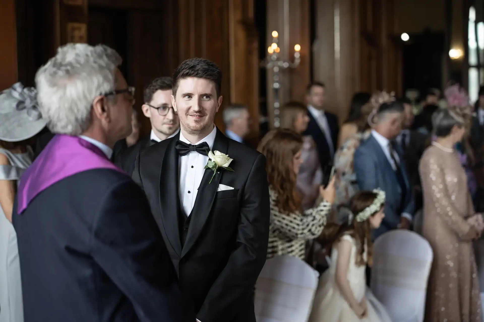 A man in a tuxedo stands facing an older man in a suit with a purple stole. Guests, including women and children, are seated in the background.