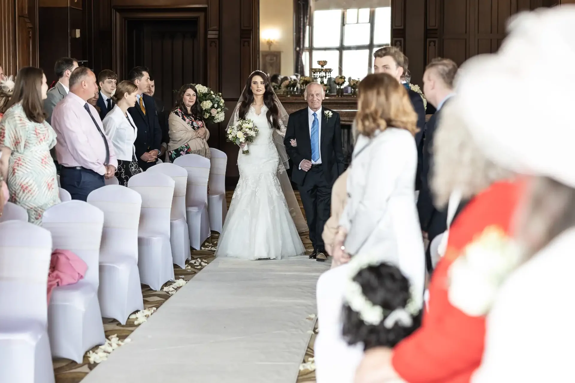 A bride walks down the aisle in a wedding ceremony, accompanied by an older man. Guests are seated on both sides, observing the procession in a decorated hall.