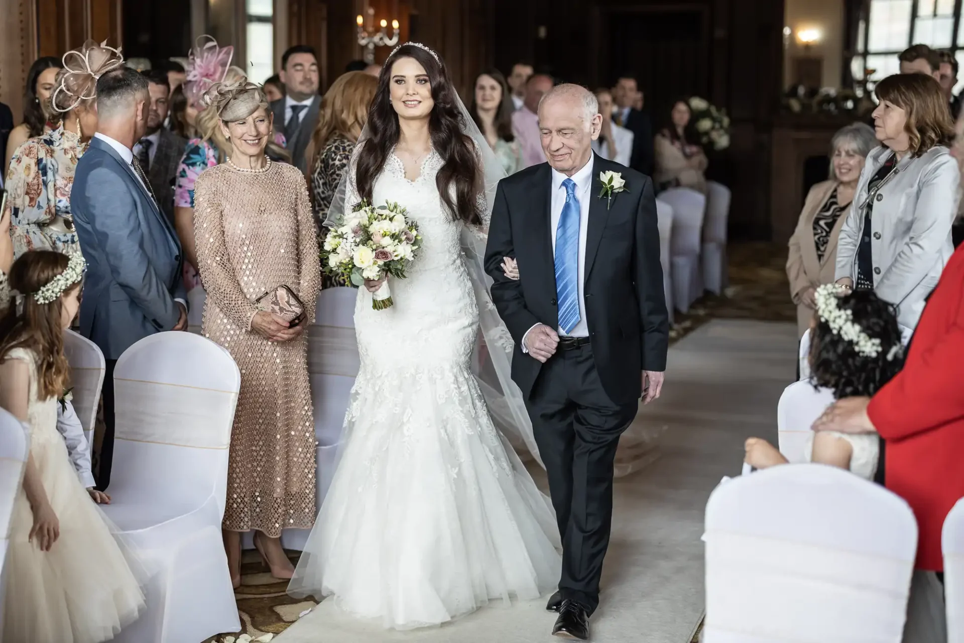 A bride in a white gown walks down the aisle with an older man in a suit, surrounded by seated guests. She holds a bouquet of flowers.