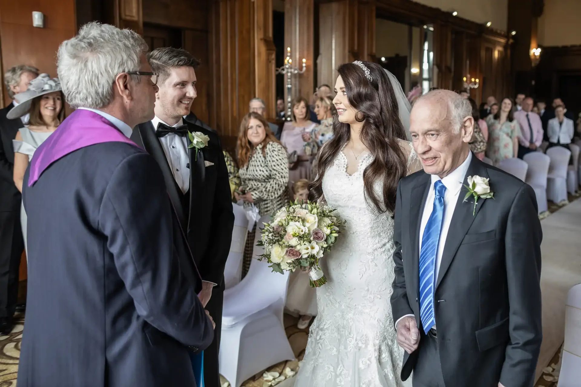 A bride in a white gown stands beside an older man, facing a groom in a tuxedo and another man in a purple stole, in a decorated room with seated guests.