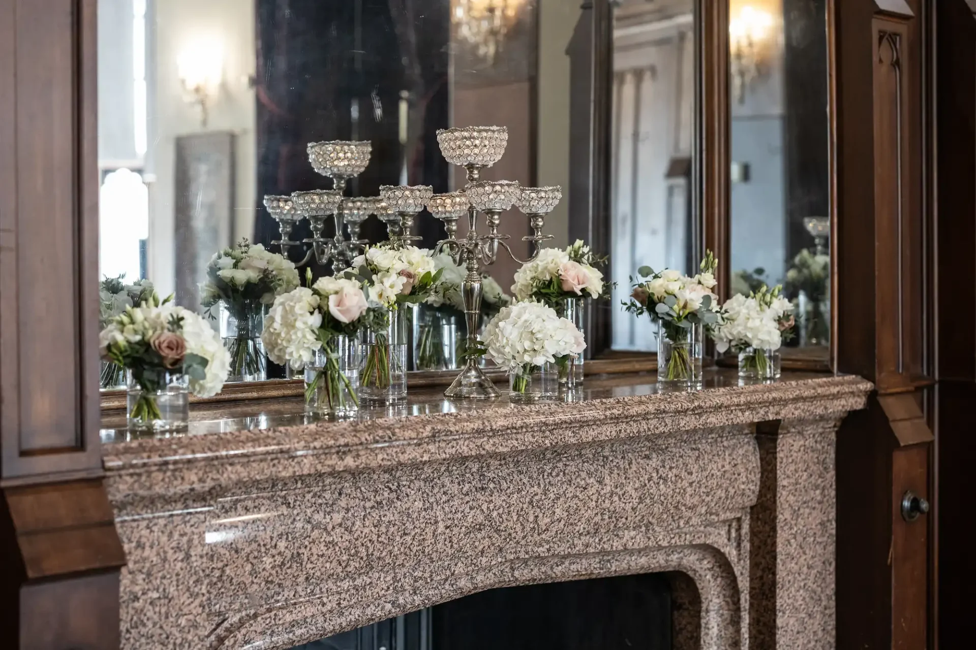 A marble fireplace mantel with a large mirror above, adorned with crystal candle holders and vases containing white and light pink flowers.