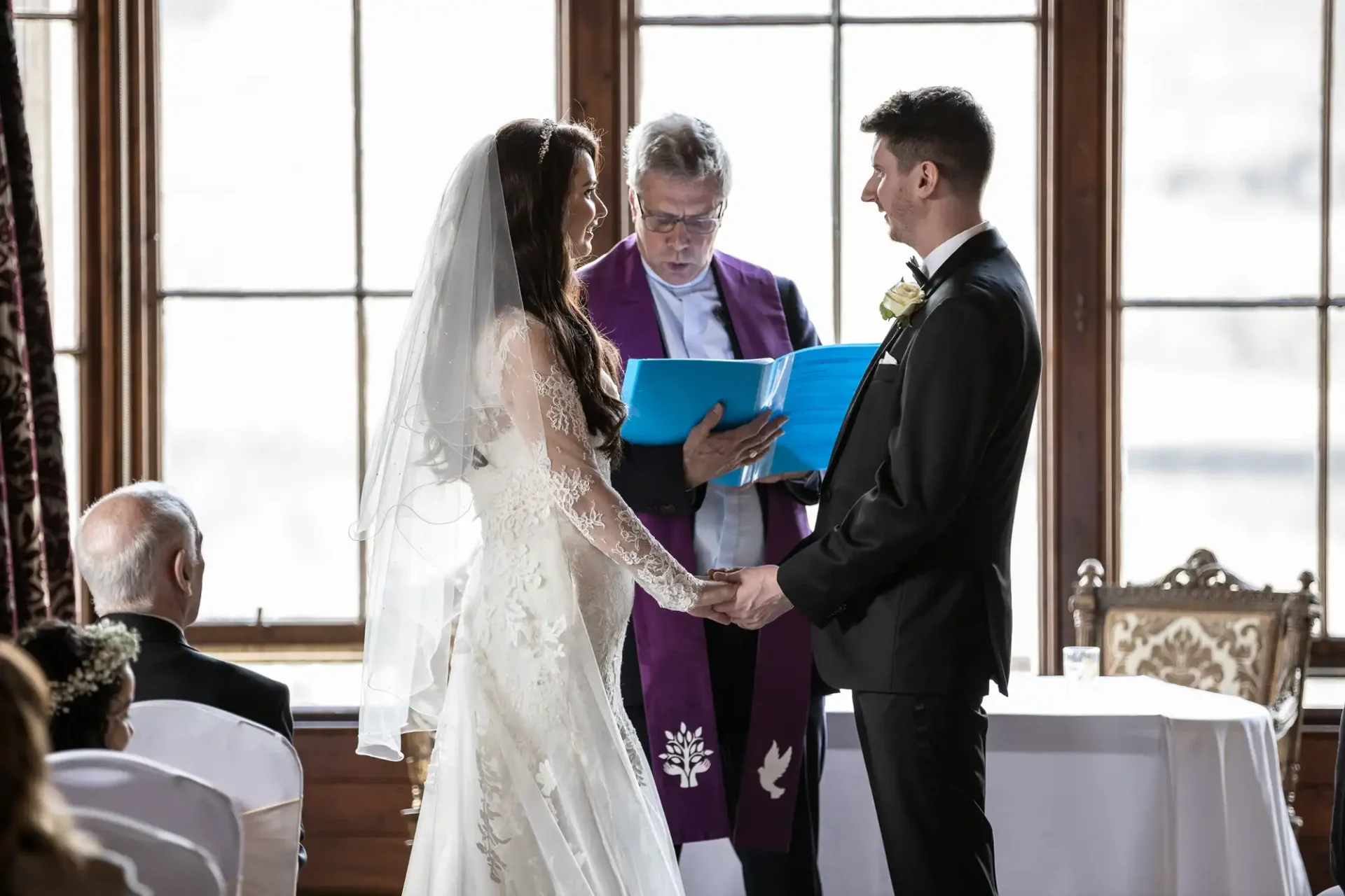 A bride and groom hold hands facing each other during a wedding ceremony, with an officiant reading from a booklet in the background.