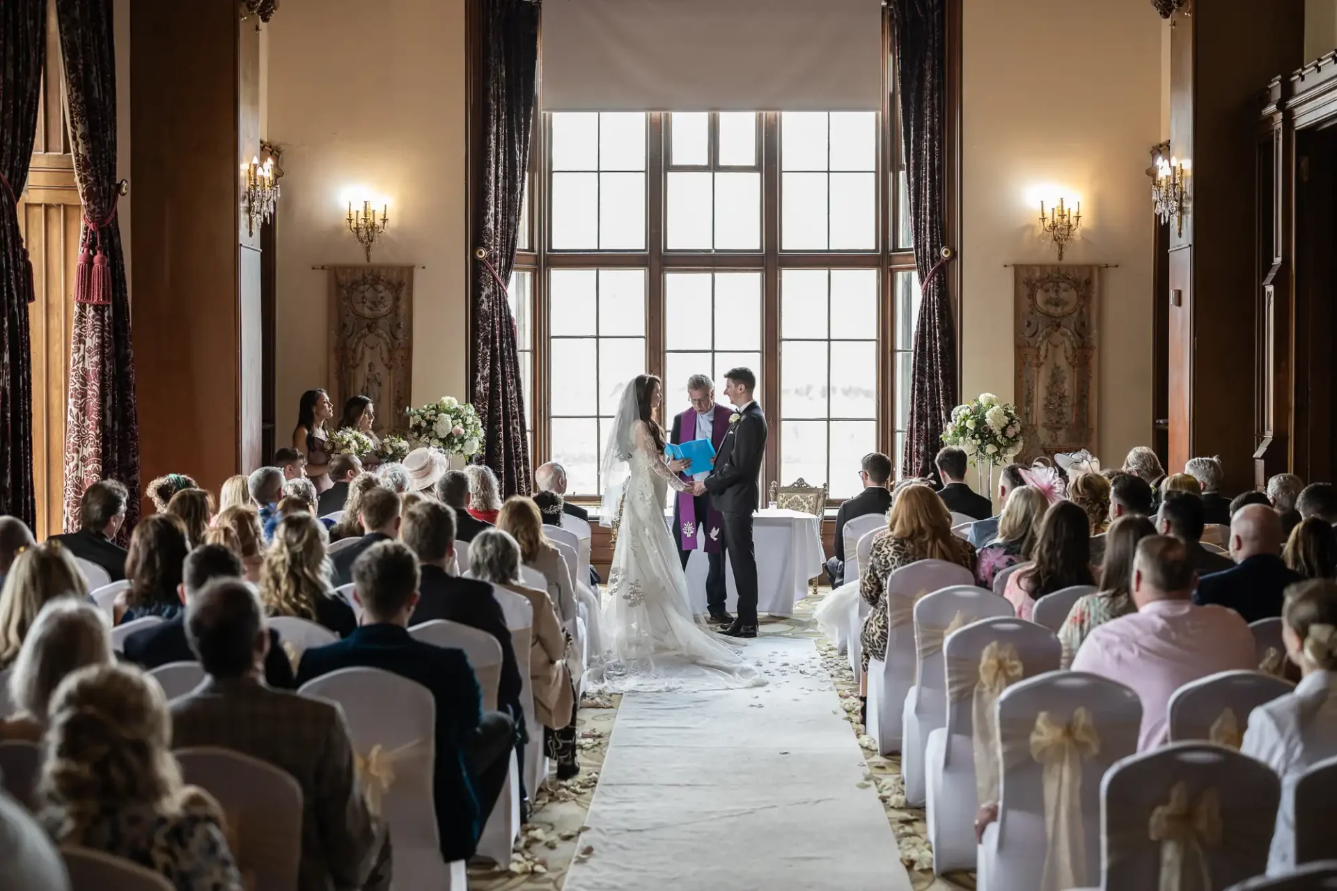 A couple stands before an officiant during a wedding ceremony in a decorated room filled with seated guests.