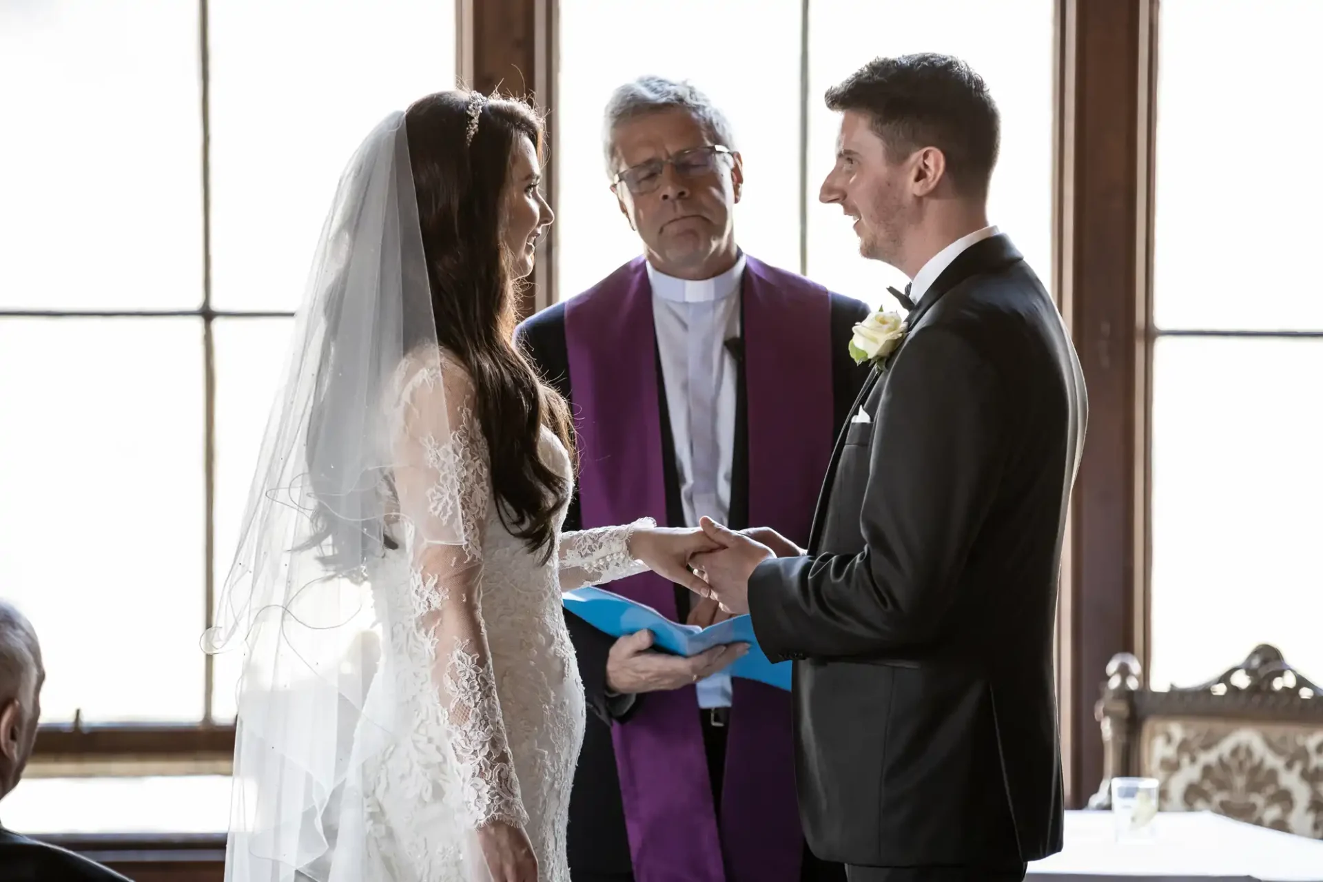 A bride and groom exchange vows holding hands during a wedding ceremony. A clergyman stands behind them holding a book.