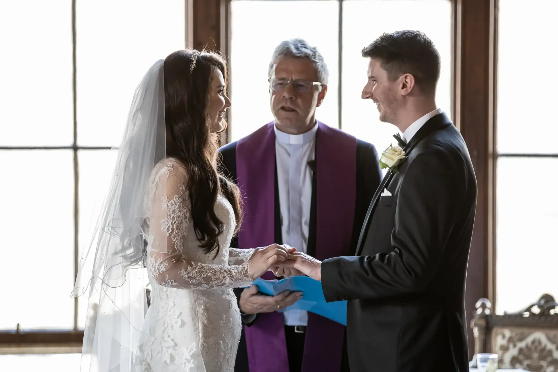 A bride and groom exchange rings during a wedding ceremony officiated by a man in a purple stole, in front of large windows.