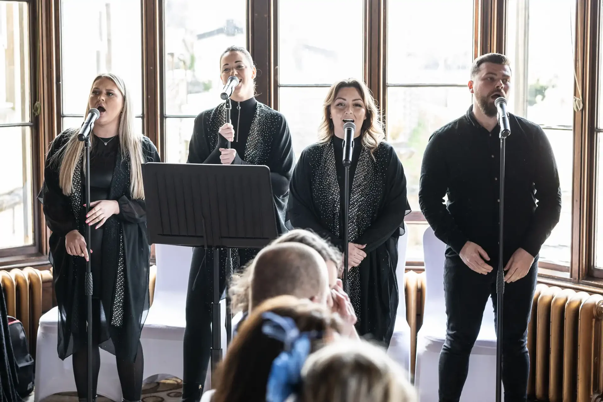 Four singers dressed in black perform with microphones in front of a window.