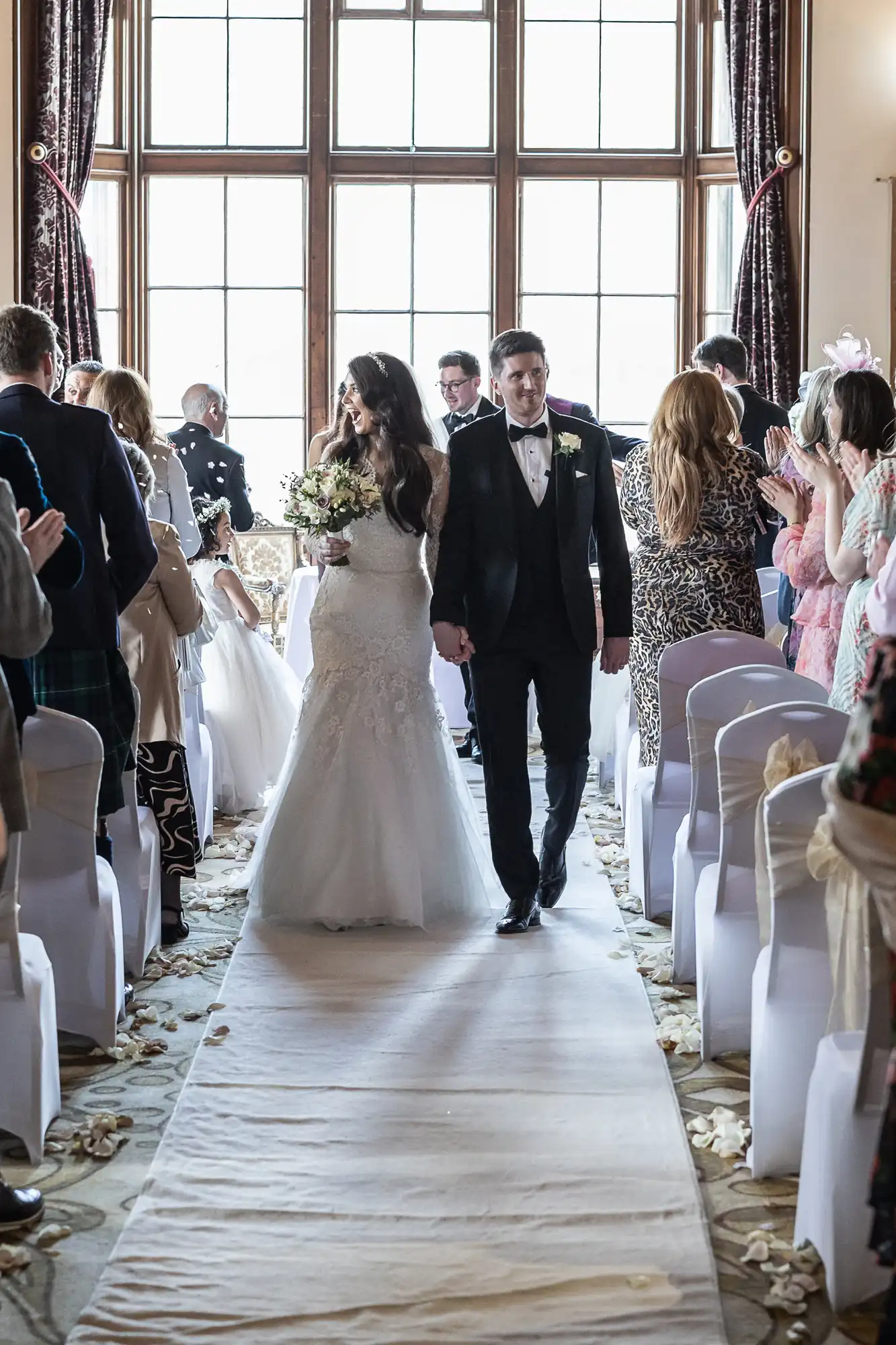 Bride and groom walk down the aisle together, surrounded by guests in a decorated venue with large windows in the background.