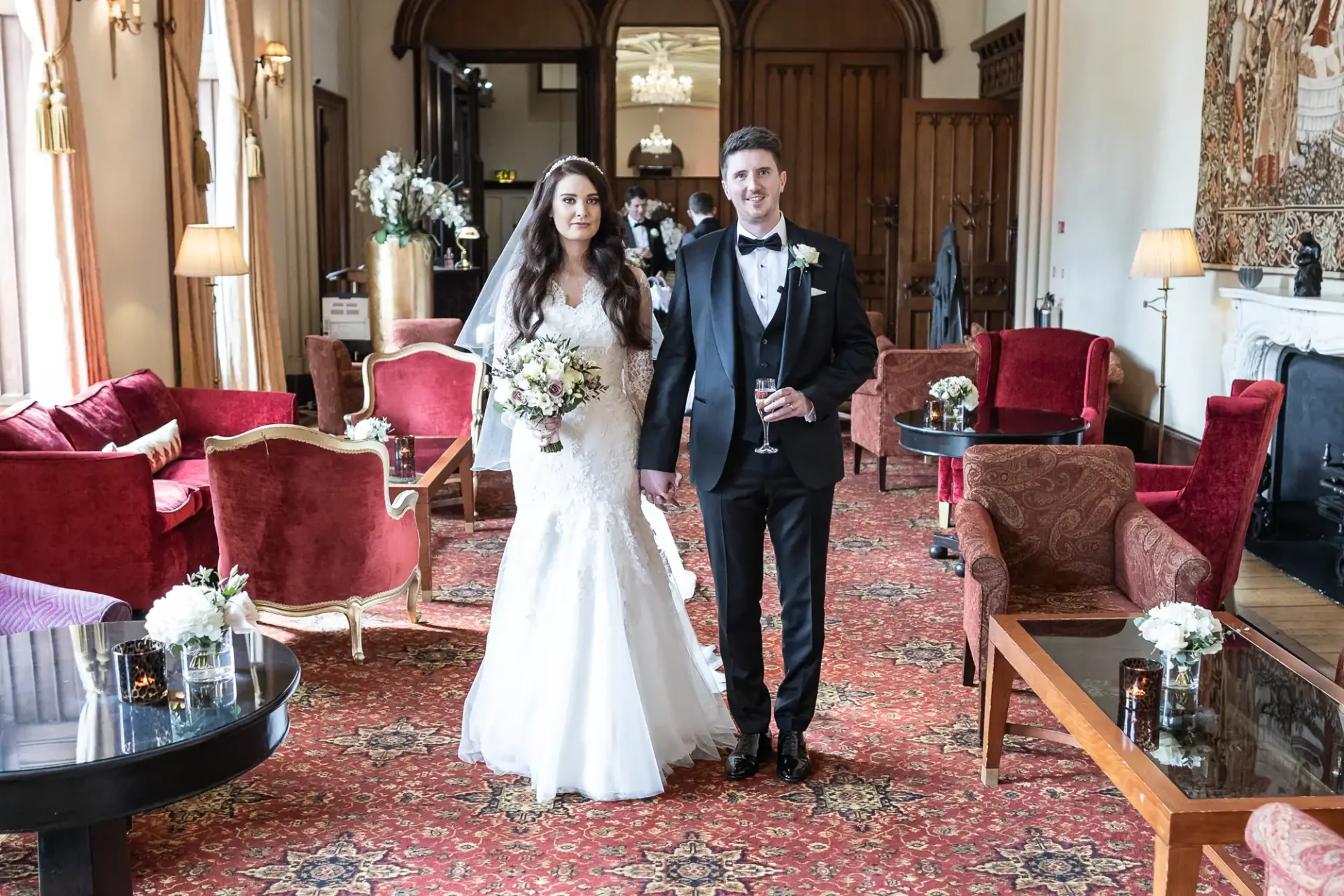 A bride and groom walk hand in hand in a elegantly decorated room with red chairs and a patterned carpet. The bride holds a bouquet; the groom holds a glass.