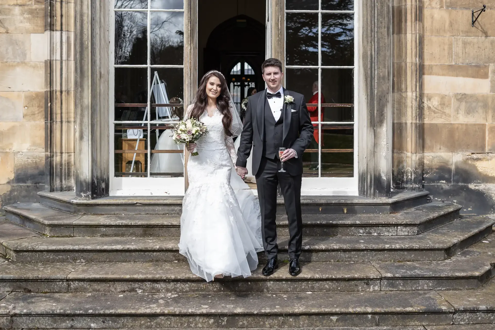 A bride in a white gown and a groom in a black suit stand together on stone steps, holding hands in front of a large windowed doorway.