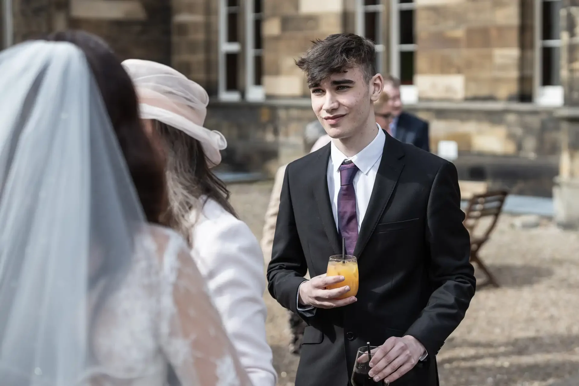 A man in a suit holds a drink and sunglasses, talking to a woman in a wedding dress outdoors.