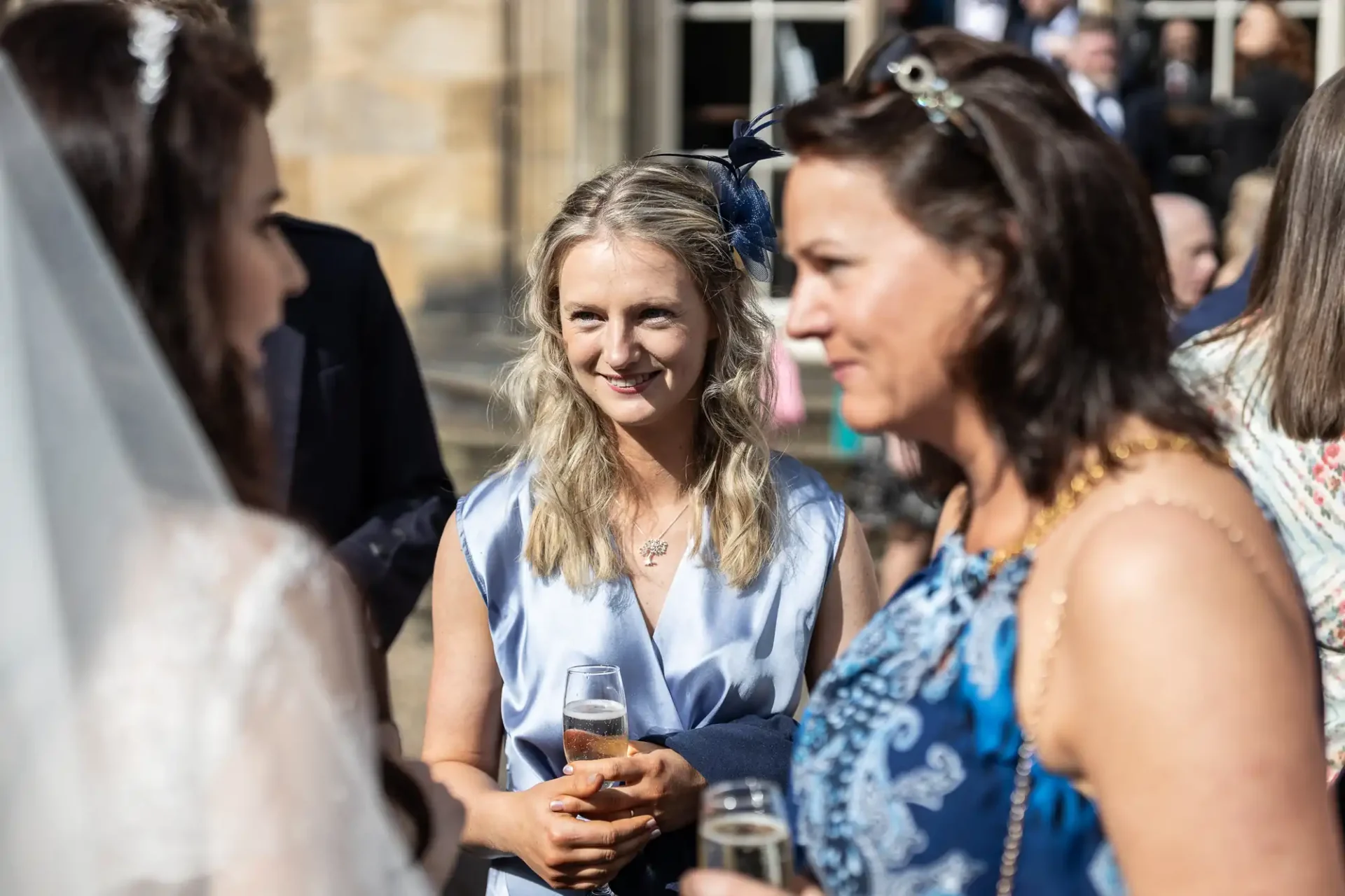 Three women in conversation at an outdoor event, two holding drinks. One wears a white veil, another a light blue dress, and the third a blue patterned dress. People are in the background.