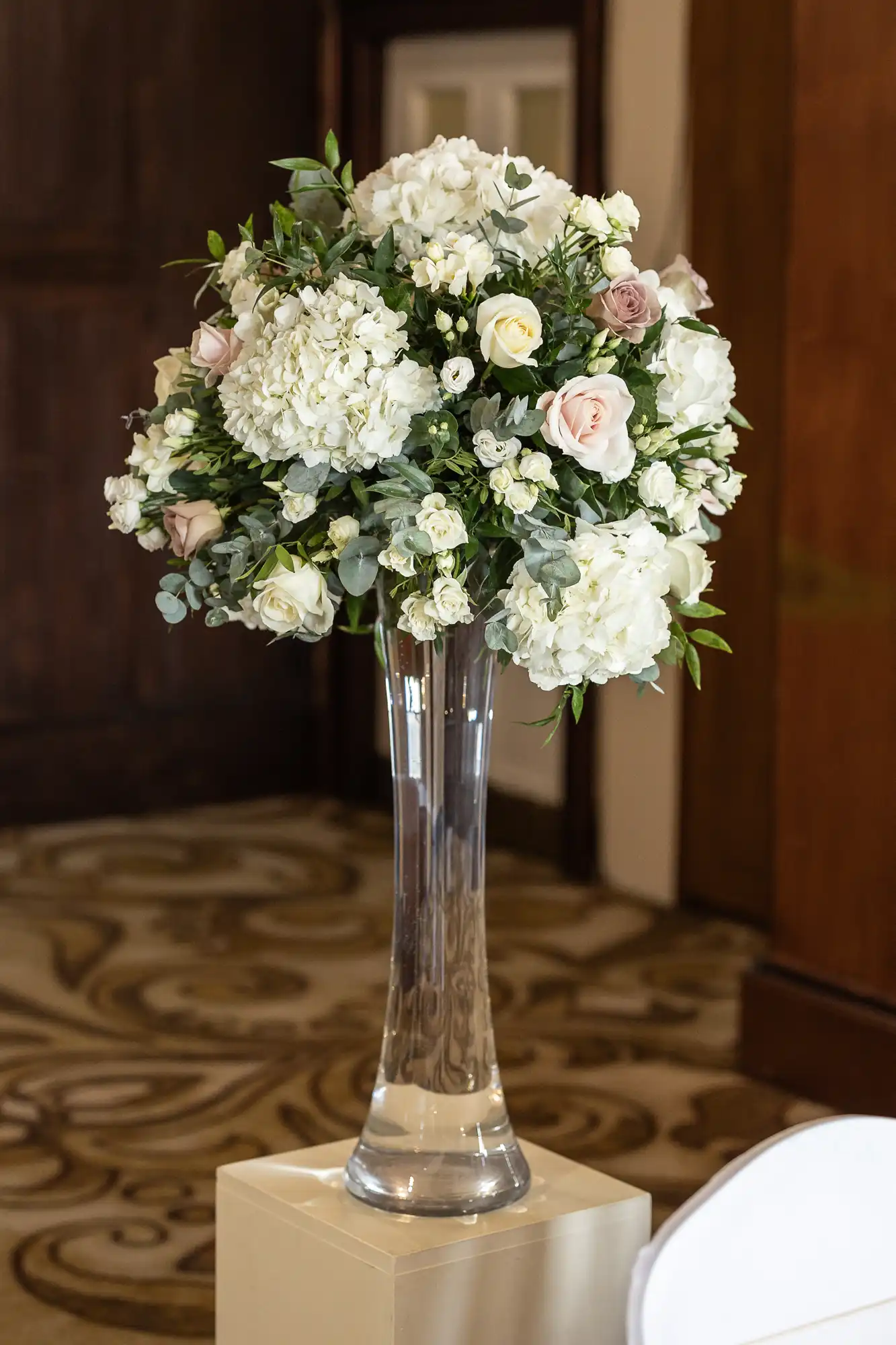 Tall glass vase with a bouquet of white and pink roses, hydrangeas, and greenery on a pedestal in an indoor setting.