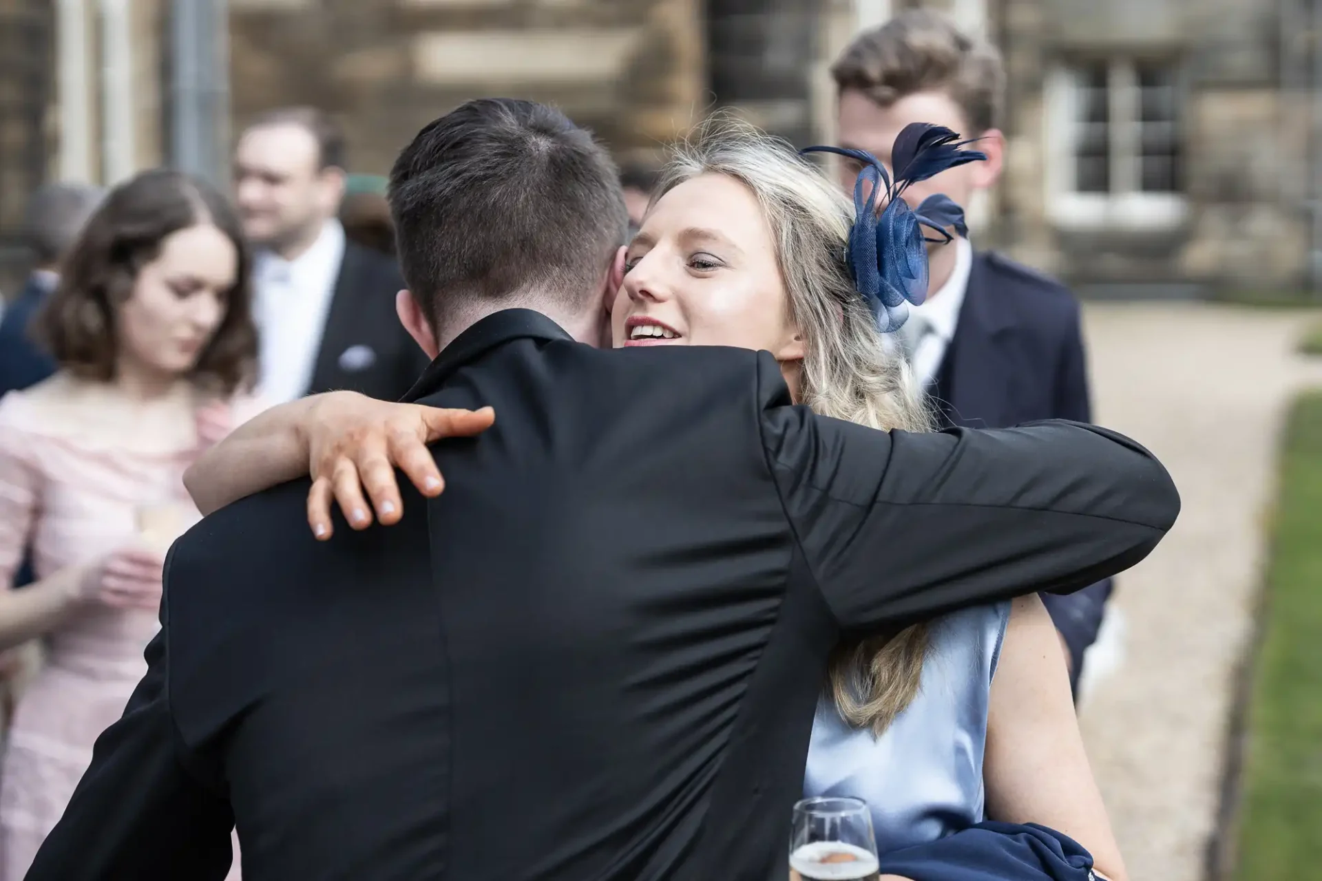 A woman in a blue dress and fascinator embraces a man in a black suit outdoors. Other people in formal attire are visible in the background.