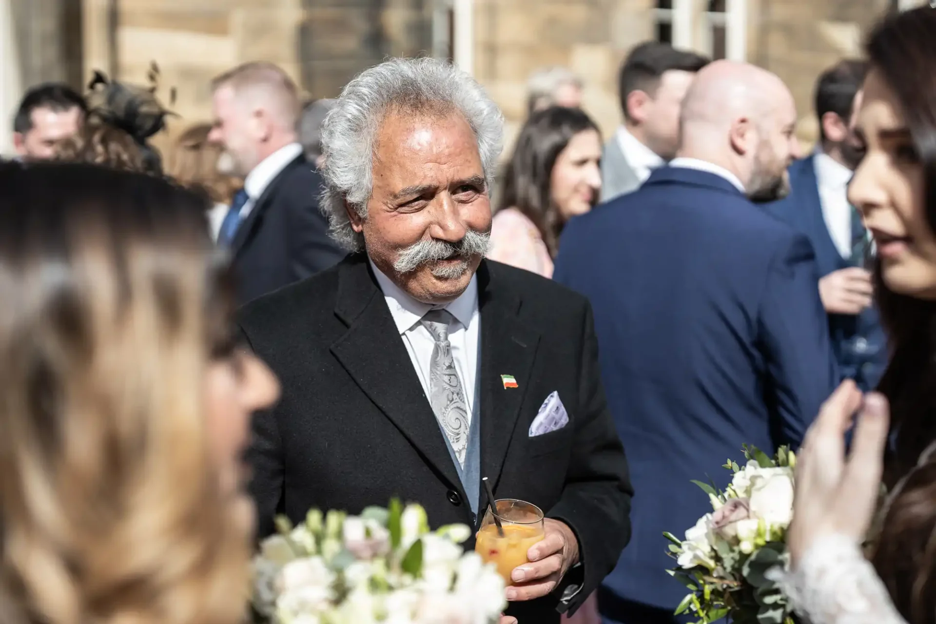 A man with white hair and a mustache in a suit, holding a drink, is surrounded by people at an outdoor gathering.