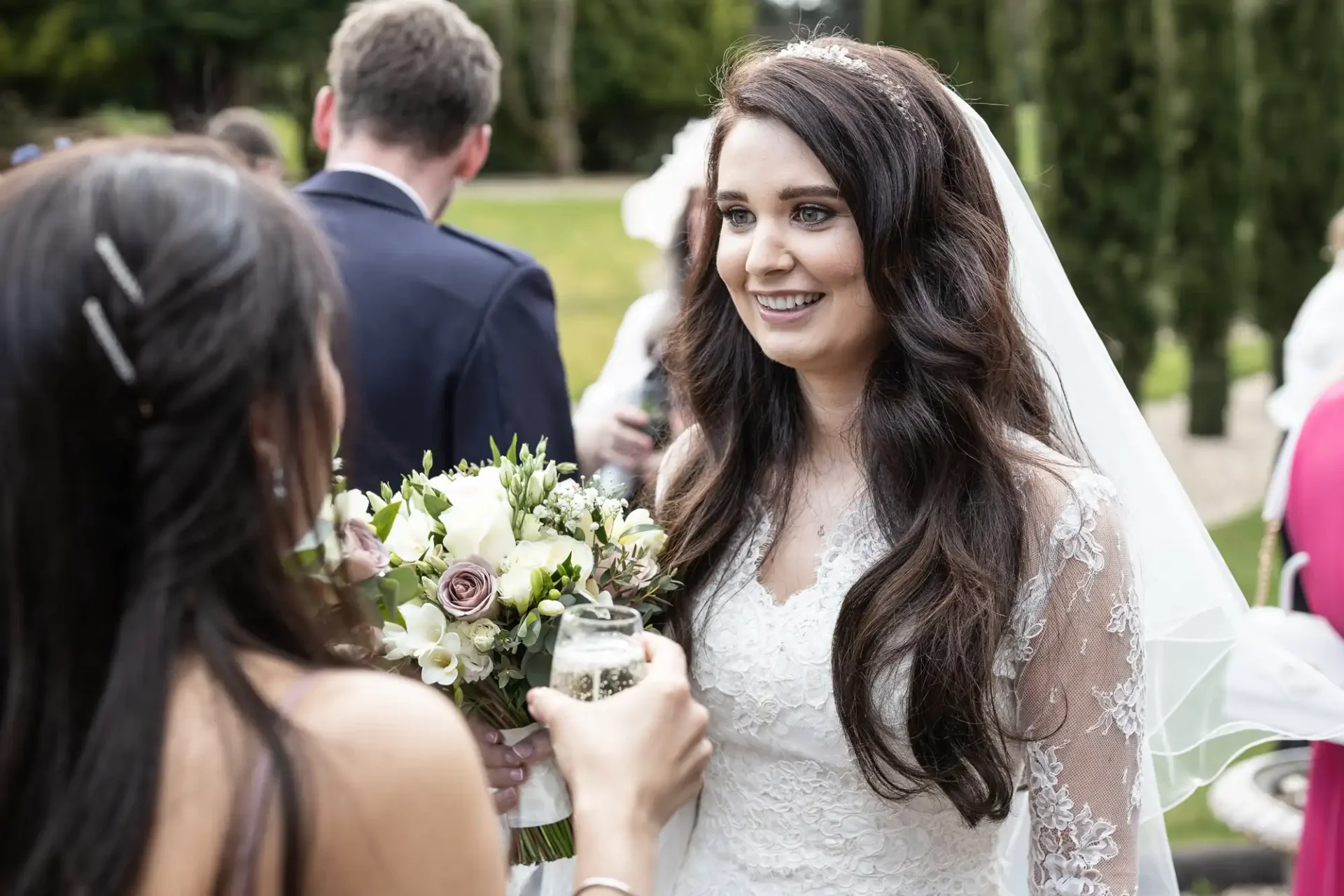 Bride smiling and holding a bouquet while talking to a guest outdoors.