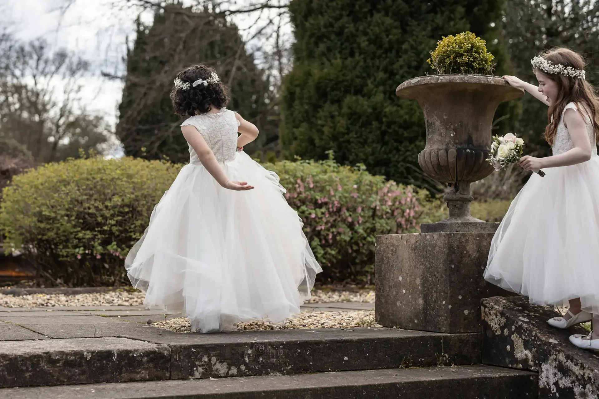 Two young girls in white dresses with flower crowns, one holding flowers, stand near a stone planter in a garden.