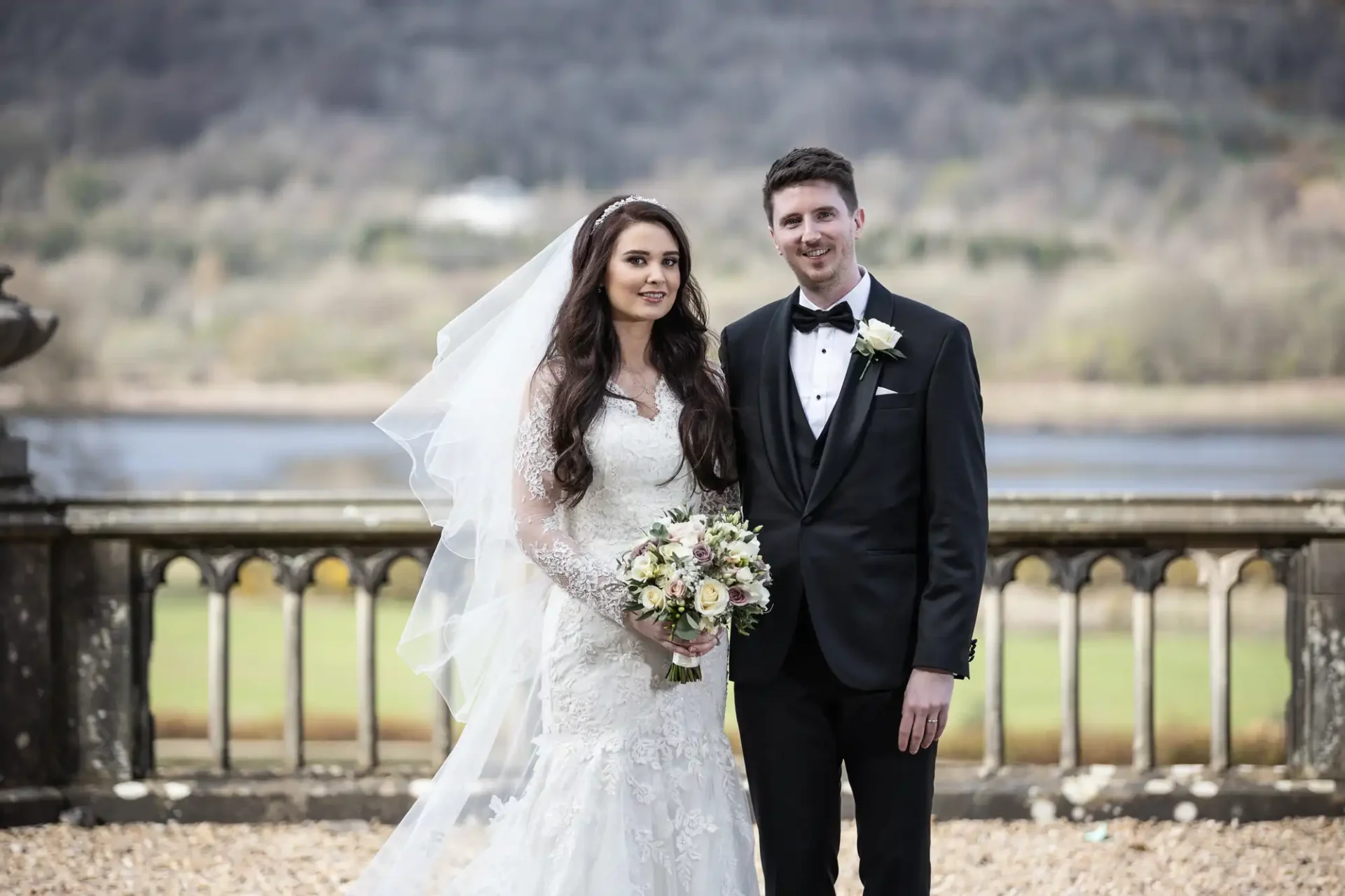 Bride in a white gown and groom in a black tuxedo stand together outdoors, with the bride holding a bouquet and a scenic backdrop of water and hills.