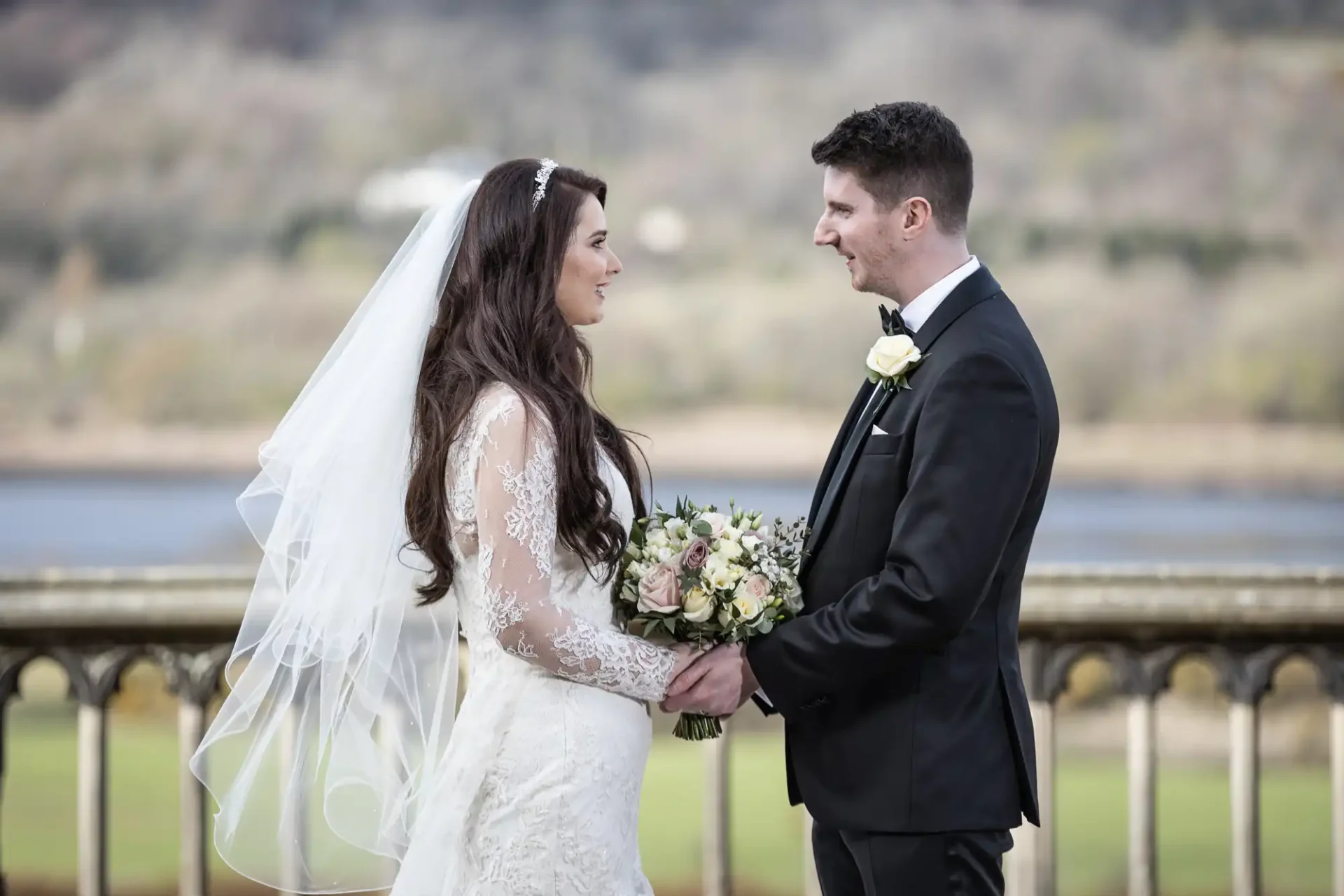 Bride and groom stand together outdoors, holding hands and smiling. Bride wears a lace gown and veil, groom is in a black suit with a white boutonniere. Scenic landscape in the background.