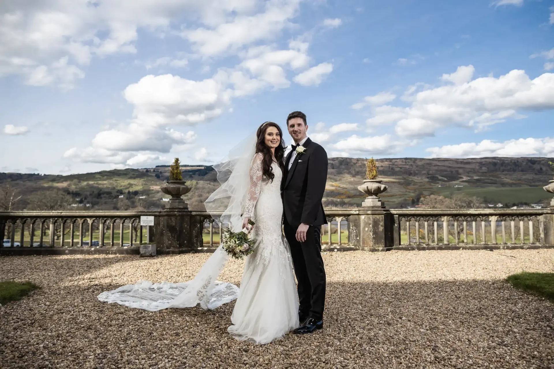 A bride and groom in wedding attire stand outdoors on a gravel path with a scenic background of hills and clouds.