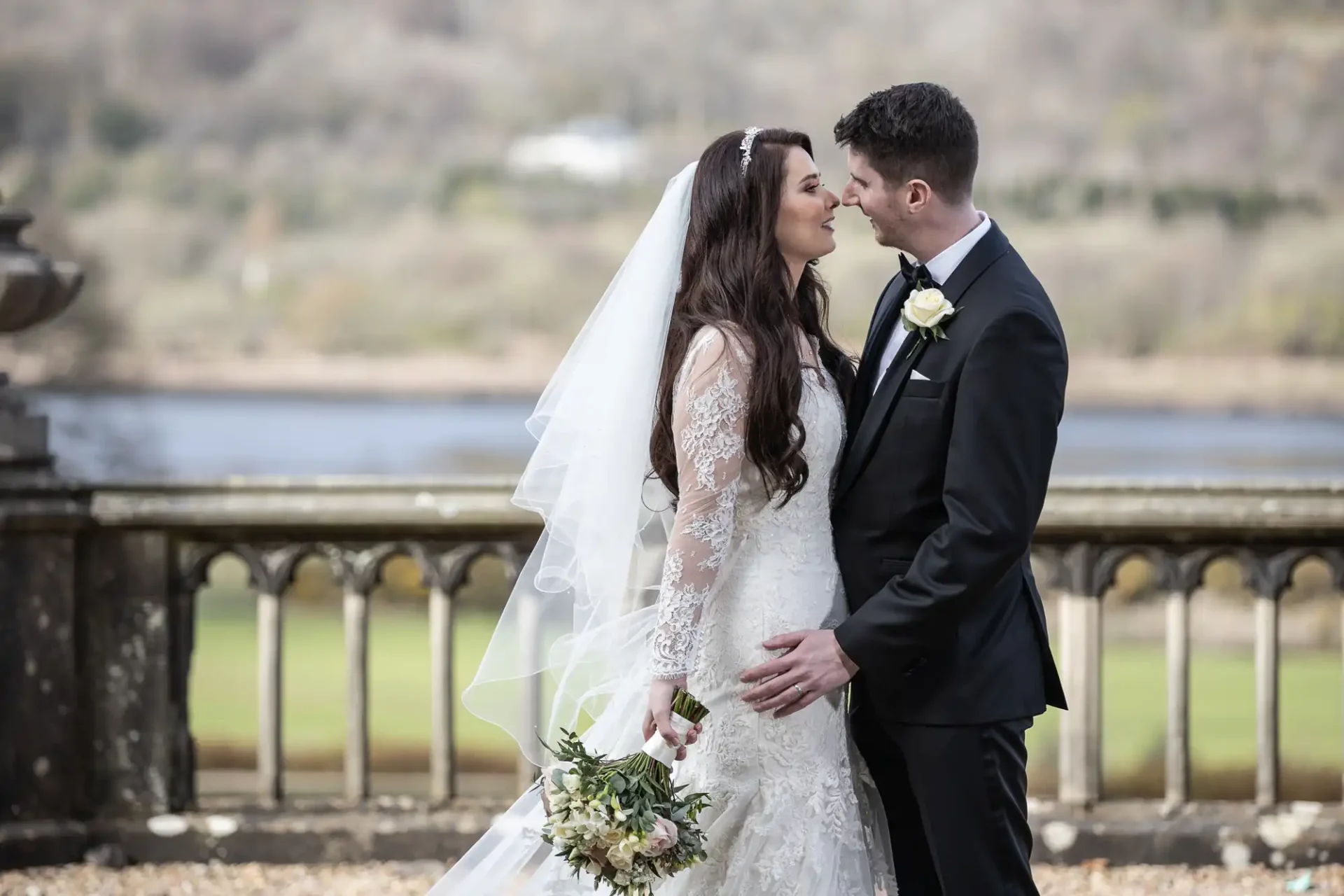 Bride and groom stand close, smiling at each other. The bride holds a bouquet, and a scenic outdoor backdrop with a lake and trees is visible.