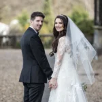 A bride and groom holding hands, standing outdoors. The groom is in a black suit, and the bride is in a lace wedding dress with a veil. Both are looking at the camera.