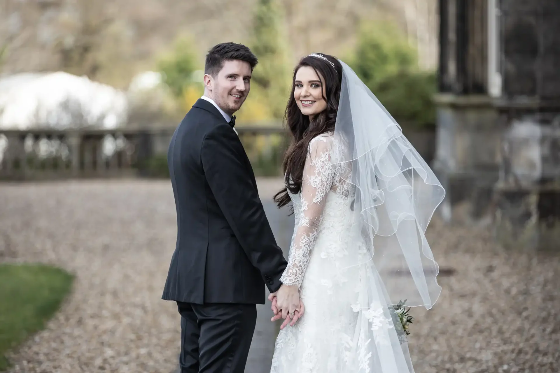 A bride and groom holding hands, standing outdoors. The groom is in a black suit, and the bride is in a lace wedding dress with a veil. Both are looking at the camera.