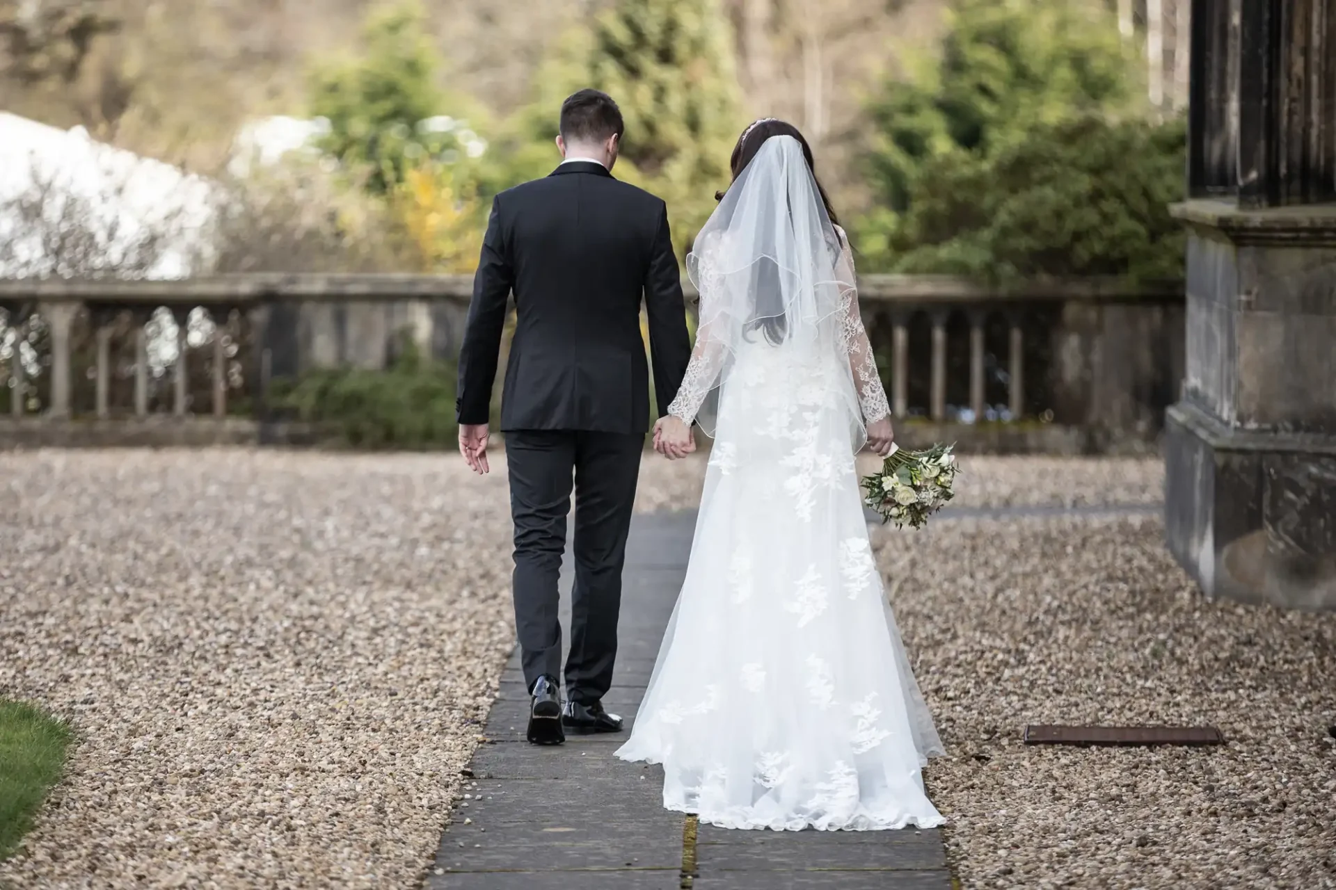 A bride and groom walk hand in hand down a stone path. The bride wears a white dress and veil, and the groom wears a black suit.