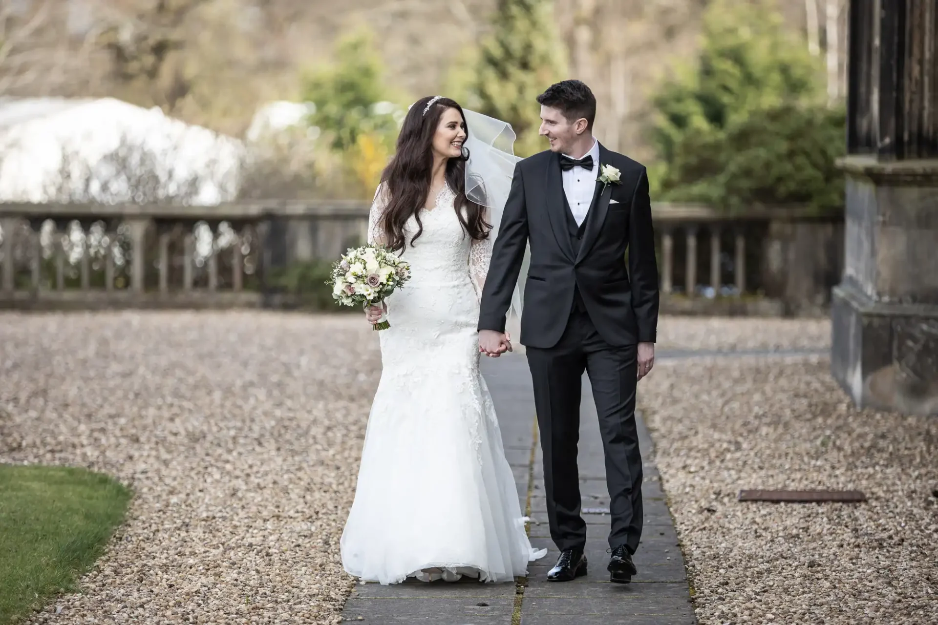 A bride in a white gown and groom in a black suit walk hand in hand outdoors on a gravel path, surrounded by greenery.