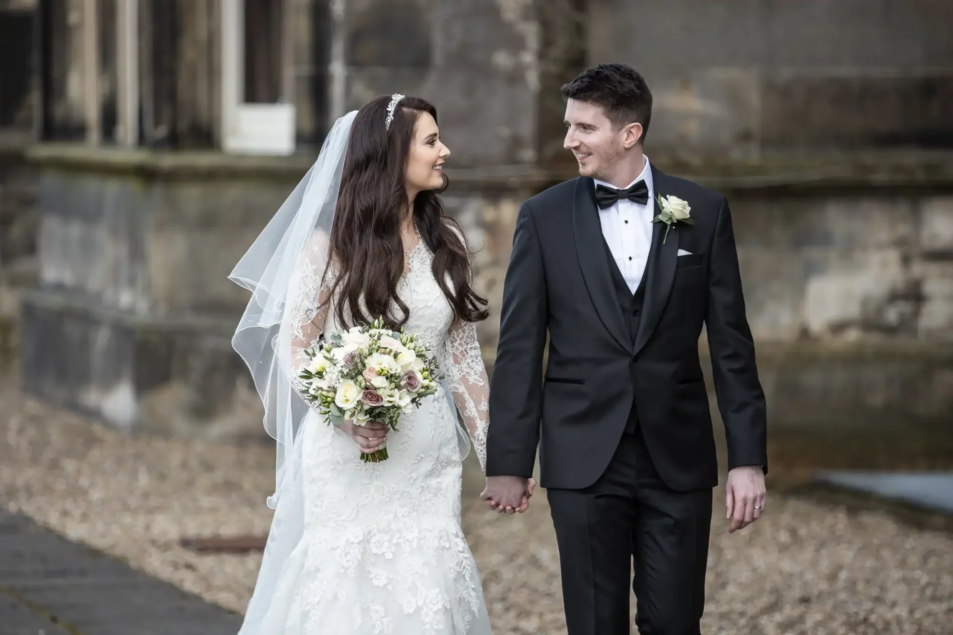 A bride in a white gown and a groom in a black suit hold hands while walking outdoors, both smiling at each other.