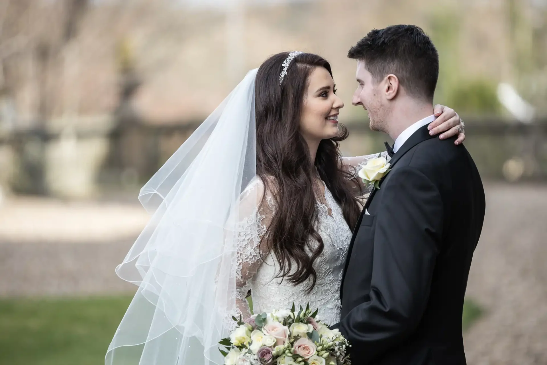 Bride in a white dress and veil smiling at groom in a black suit and bow tie, outdoors with a bouquet of flowers.