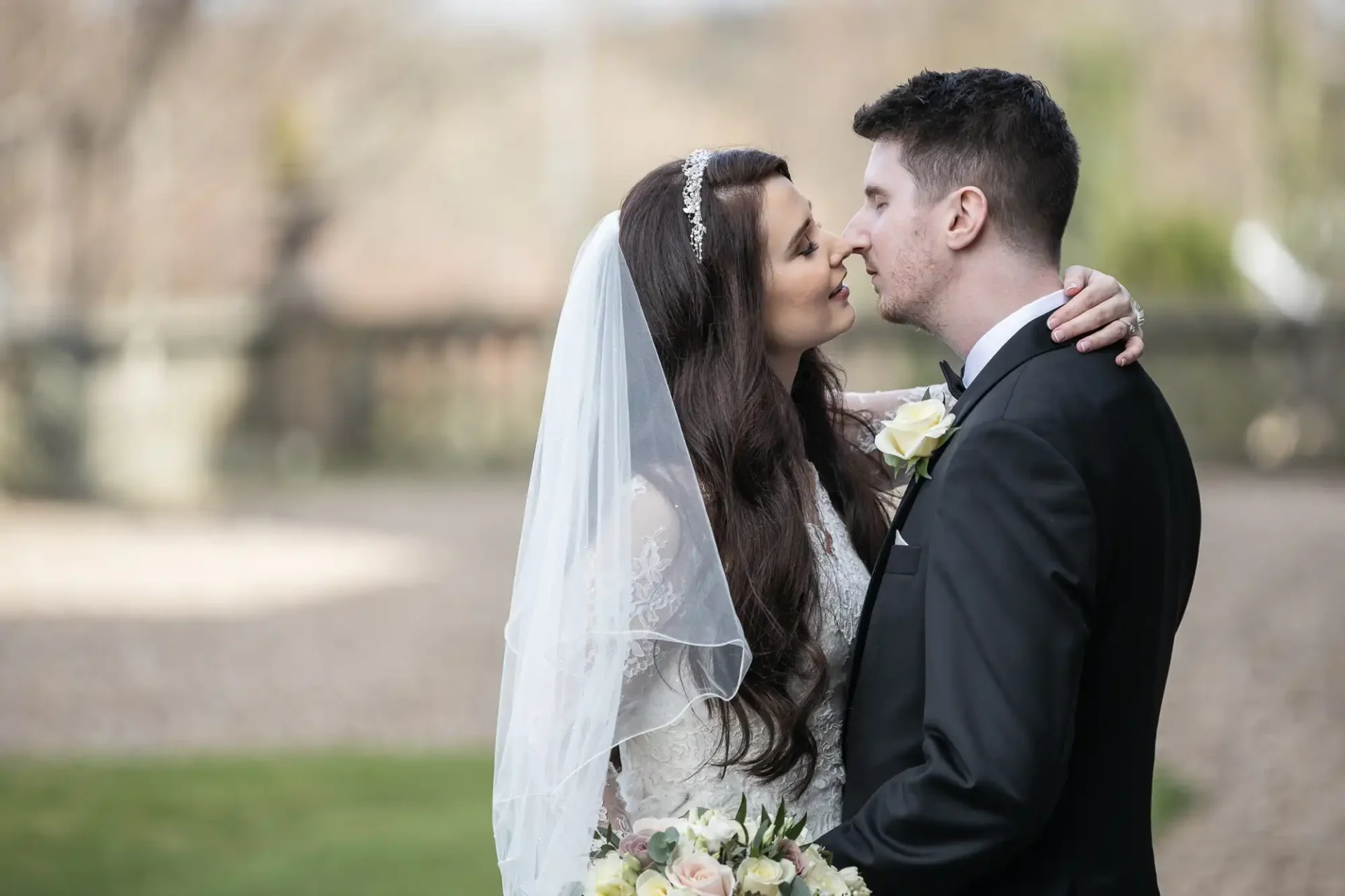 Bride and groom standing closely, about to kiss, outdoors. Bride holds a bouquet and wears a lace dress with a veil. Groom is in a black suit with a boutonniere.