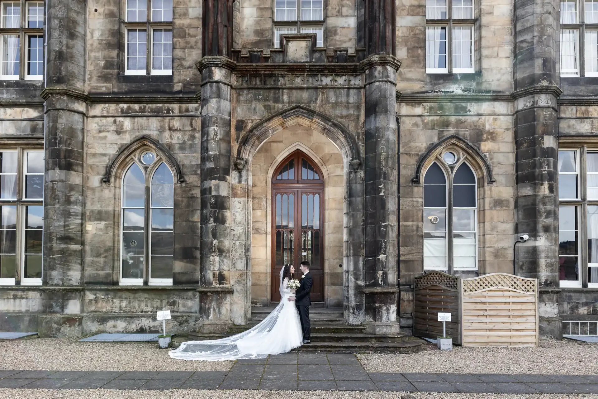 Bride in white gown and groom in black suit stand in front of a large, ornate stone building with arched windows and wooden door.