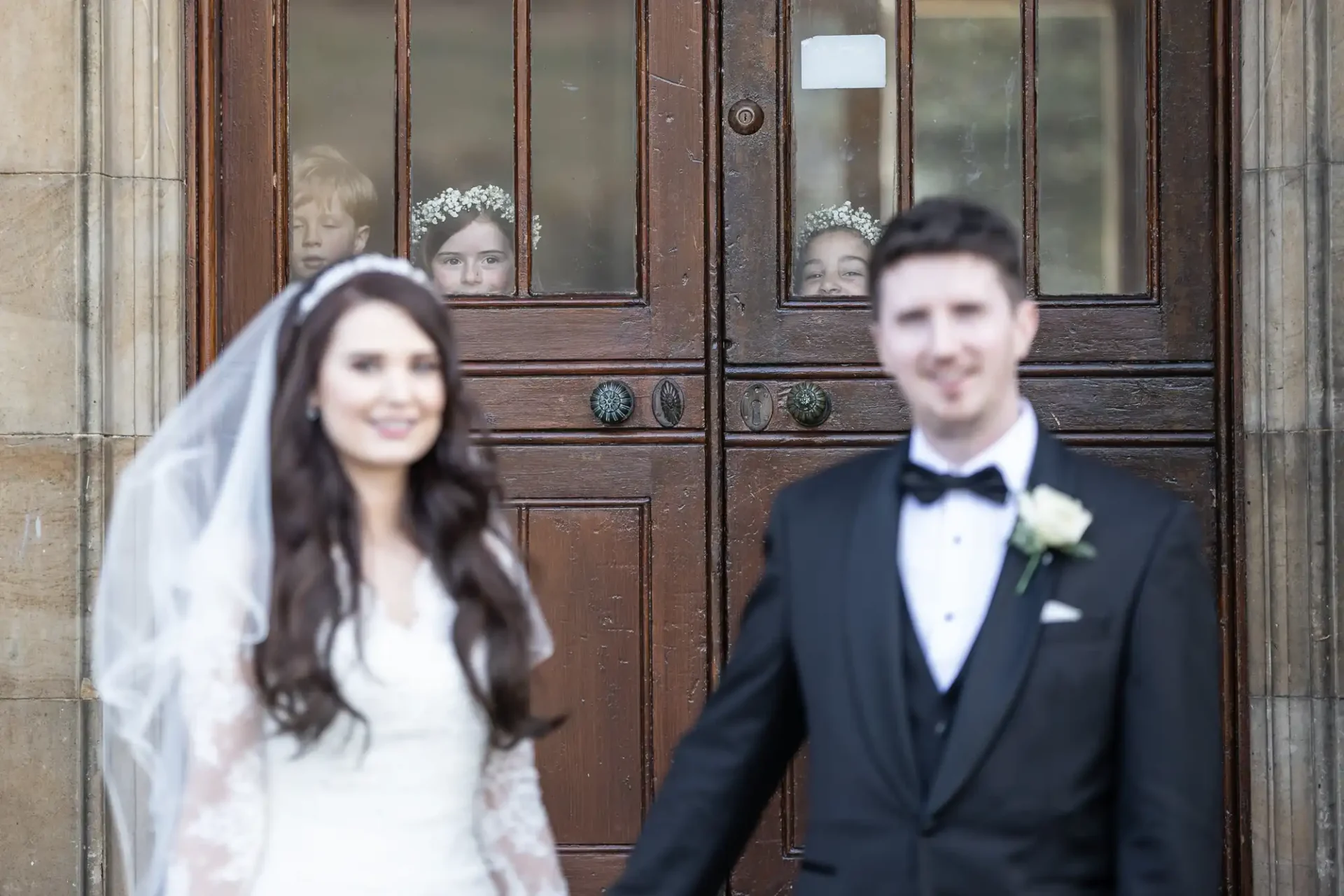 Bride and groom stand in front of a wooden door. Three children with flower crowns are visible through the door's glass panes in the background.