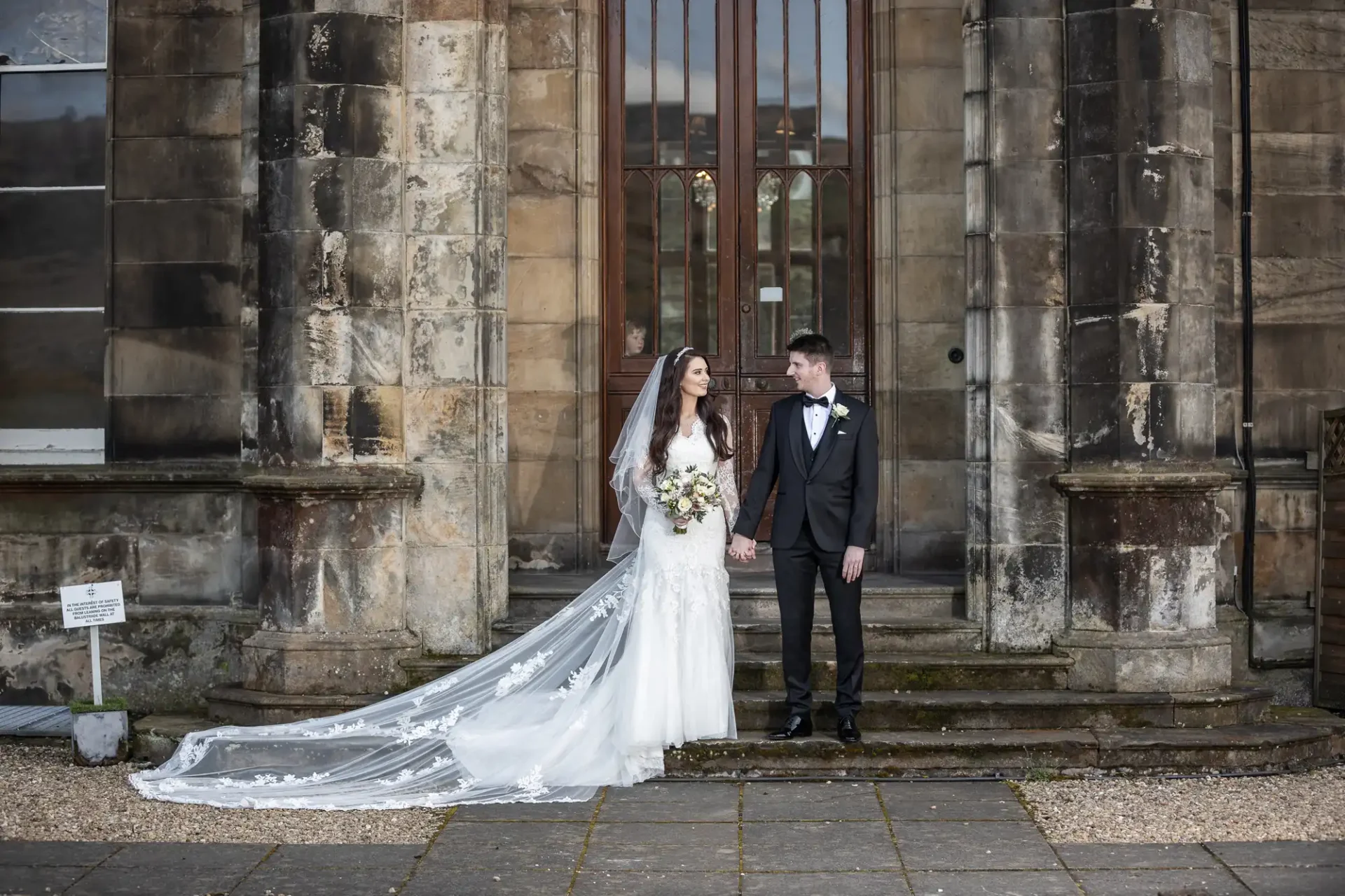 Bride and groom standing in front of a stone building. The bride is in a white gown with a long train, and the groom is in a black tuxedo. They are holding hands and looking at each other.