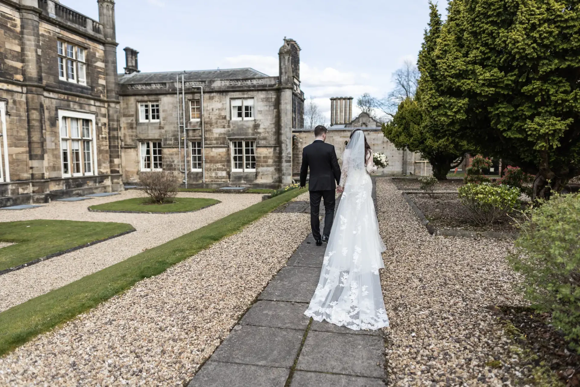 A bride and groom walk along a stone path near an old building with manicured gardens.