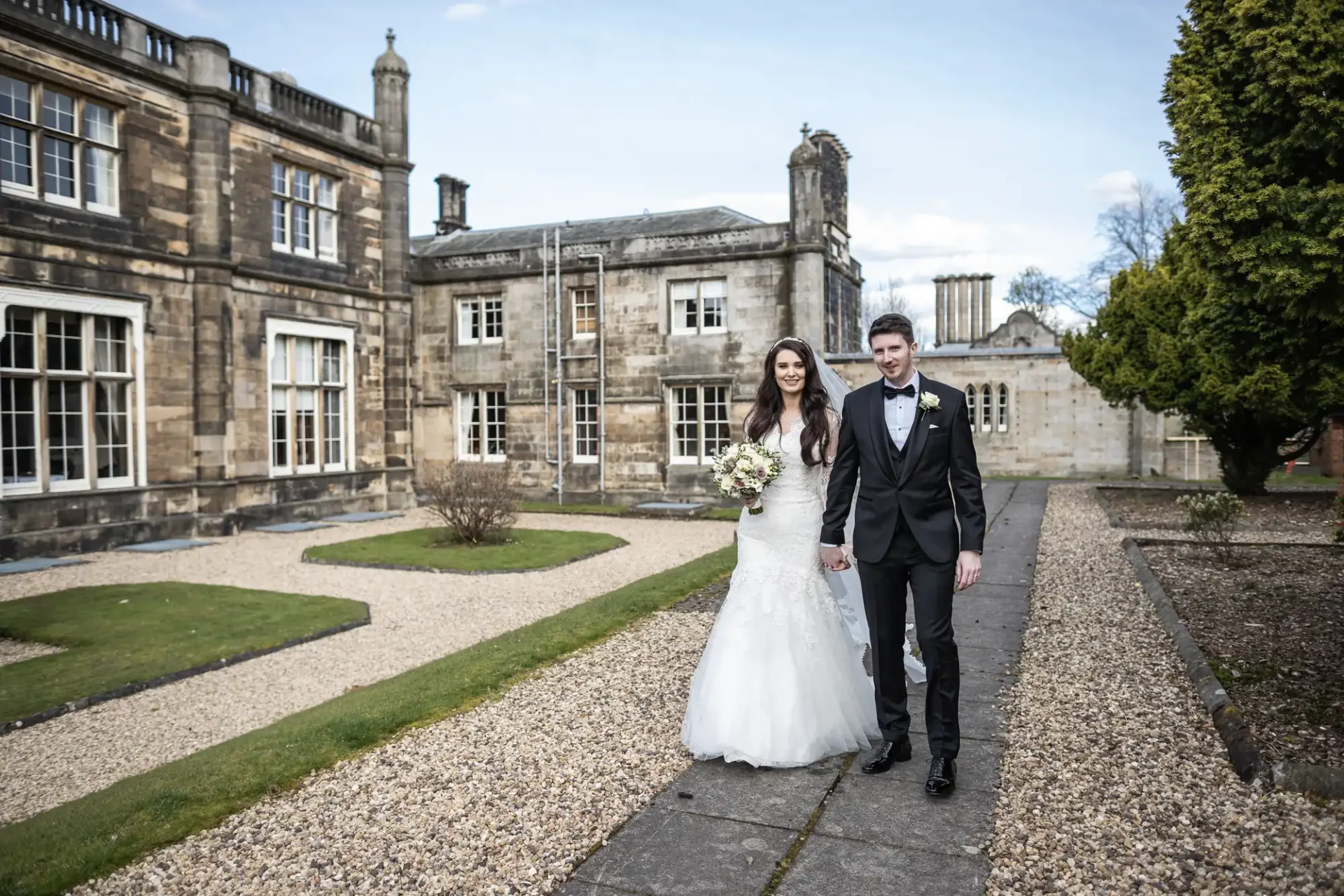 A couple in wedding attire walking on a stone path in front of a historic building. The bride holds a bouquet, and both are smiling.