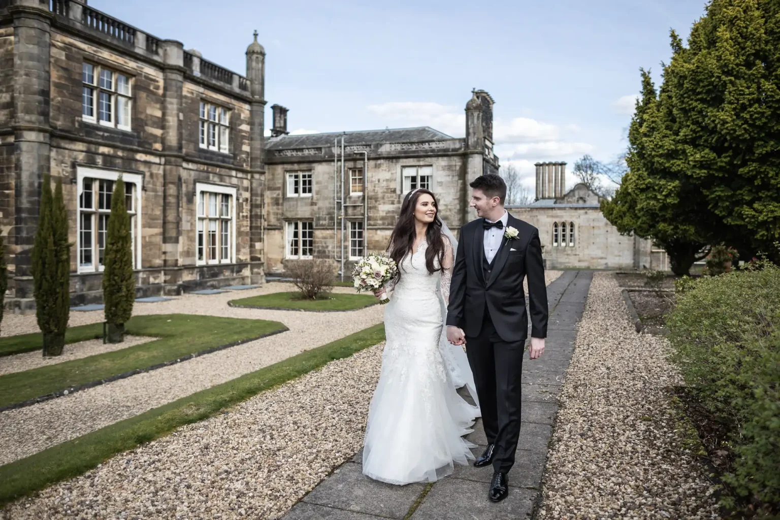 Mar Hall wedding photos: A couple in wedding attire holding hands, walking on a path in front of a historic stone building and manicured garden.
