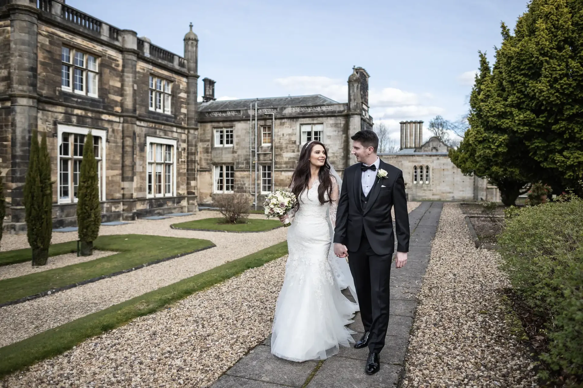 Mar Hall wedding photos: A couple in wedding attire holding hands, walking on a path in front of a historic stone building and manicured garden.