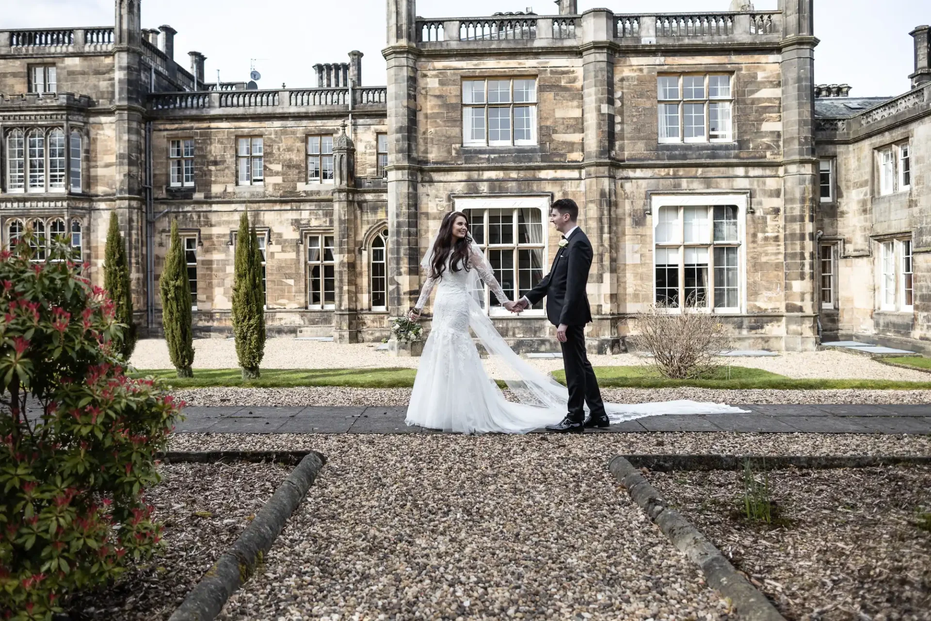 A bride and groom stand holding hands in front of a large, historic stone building with multiple windows and columns.