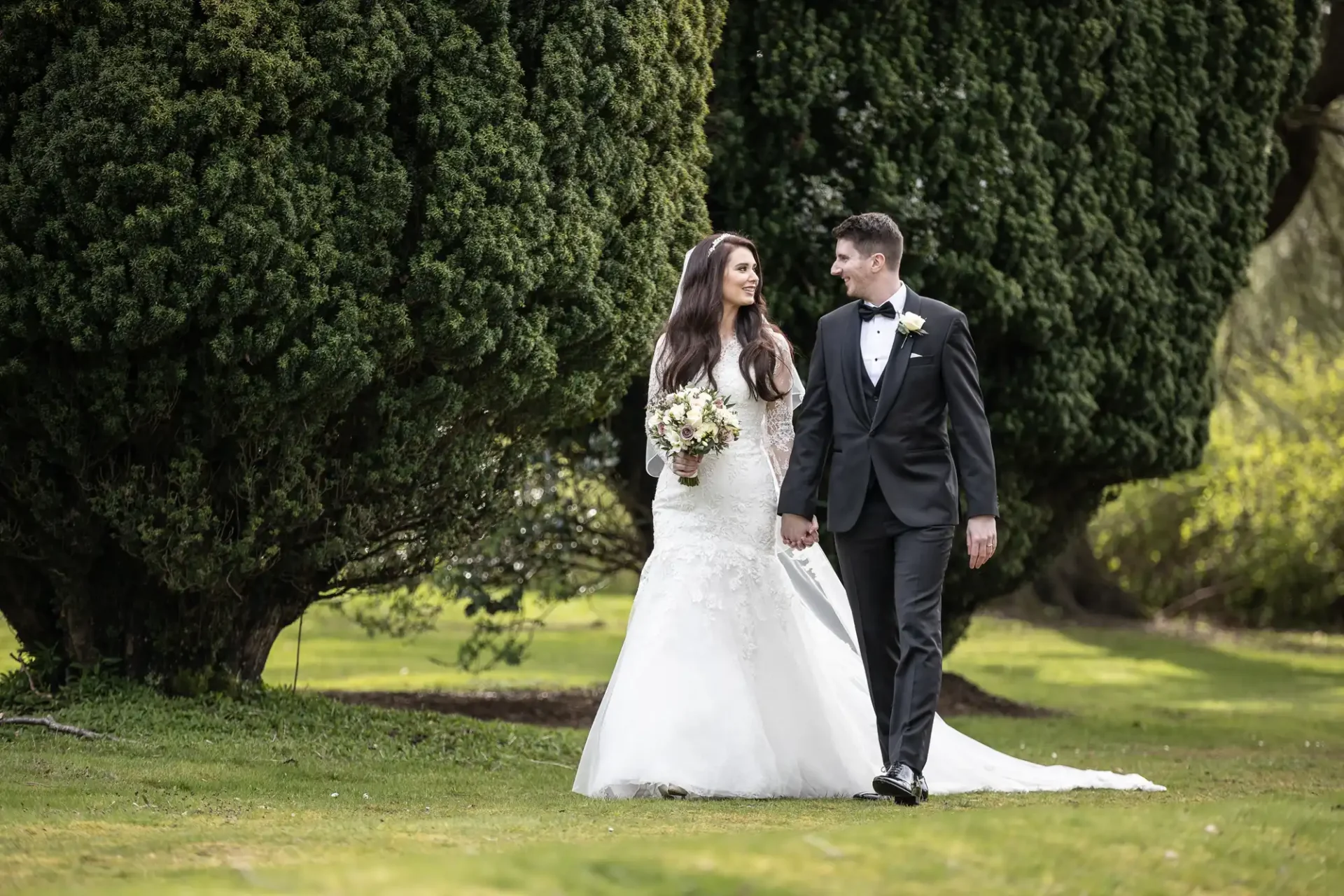 A bride in a white gown and a groom in a black suit walk hand in hand on grass, with large trees in the background.