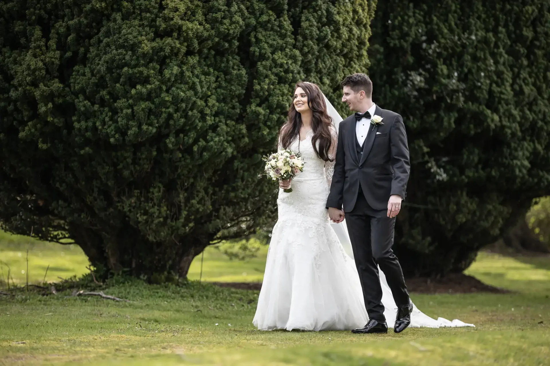 A bride in a white gown and a groom in a black suit hold hands while walking on grass, with large trees in the background.