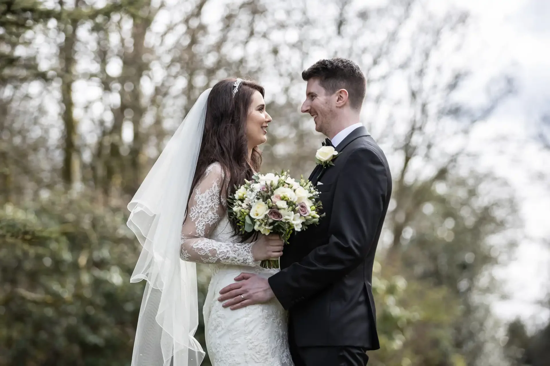 Bride and groom standing outdoors, facing each other. The bride holds a bouquet, and both are smiling. Trees are visible in the background.