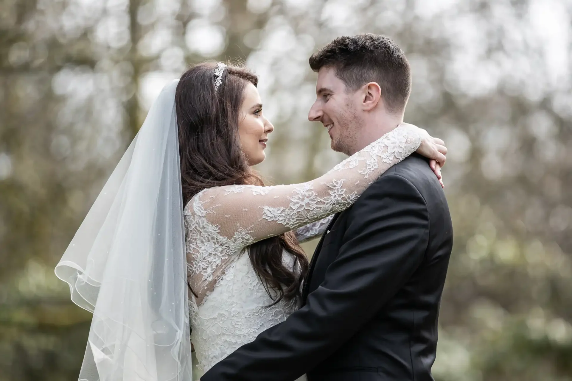 A bride and groom embrace outdoors, gazing at each other. The woman wears a white lace dress and veil, and the man is in a black suit. The background is blurred greenery.