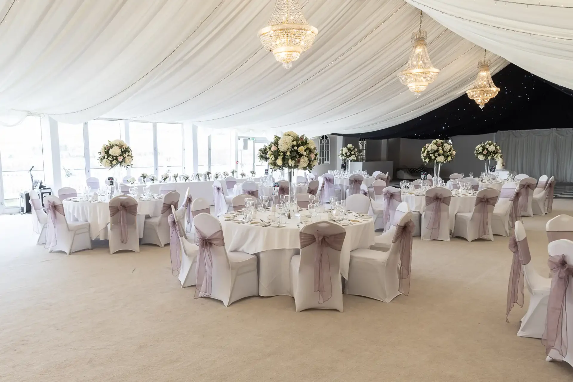 A wedding reception venue with round tables, white tablecloths, and chairs adorned with lavender sashes. Chandeliers hang from a draped ceiling.